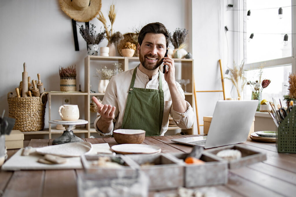 small business owner talking on a mobile phone with a customer with various creative art projects on the desk in the foreground