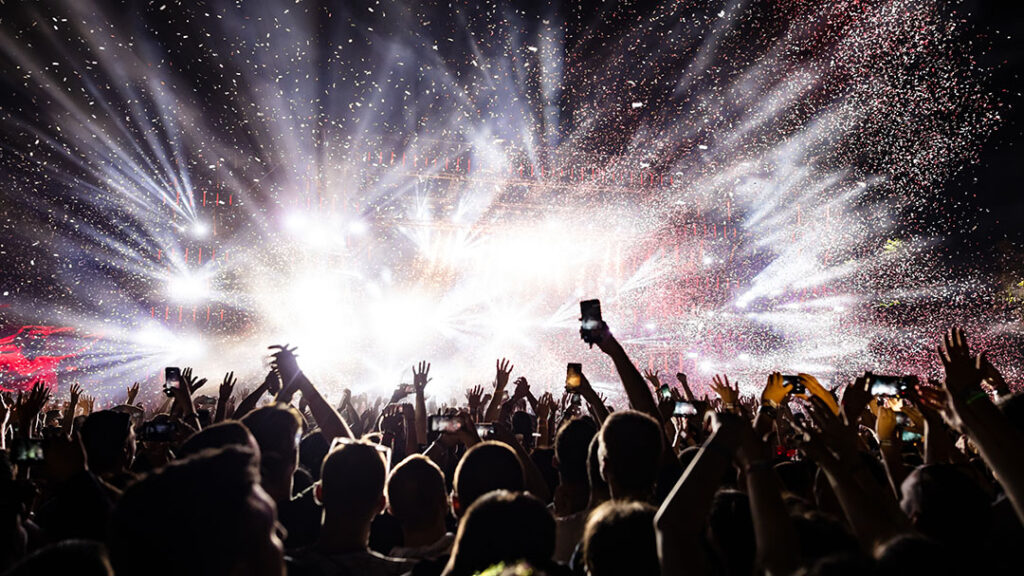 View of a concert from the back of an arena with fans holding cell phones up and an unrecognizable act on stage represented by bright lights