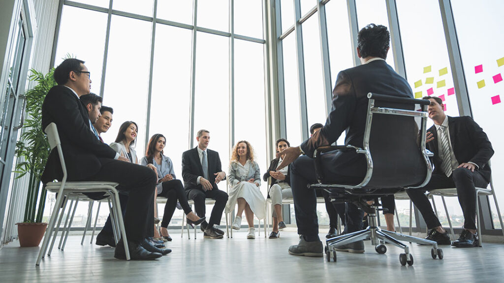 Group of business professionals sitting in a circle in a large high ceiling conference room with large windows and natural lighting