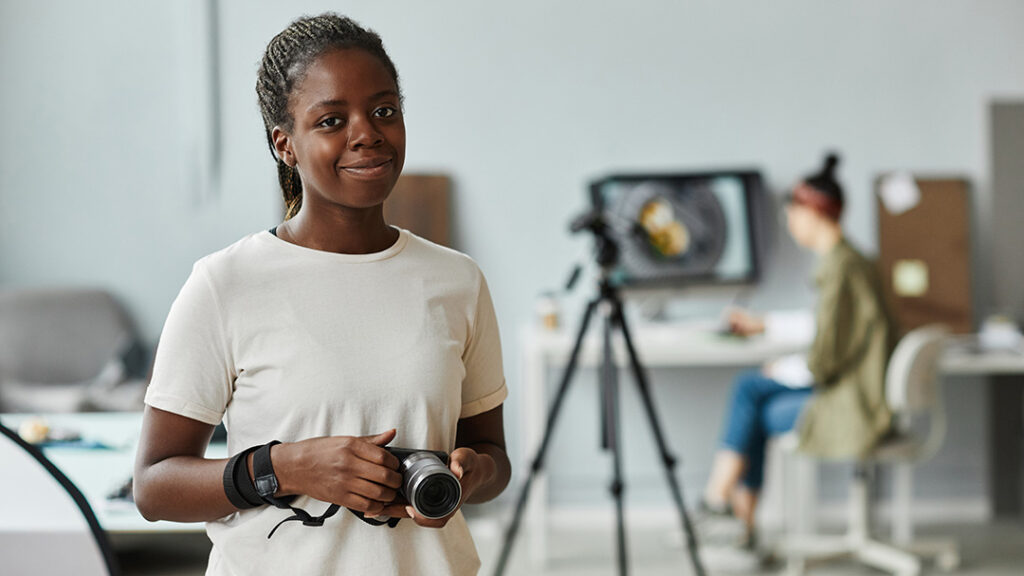 female photographer smiling with a subject to be photograph in the background behind a camera with tripod