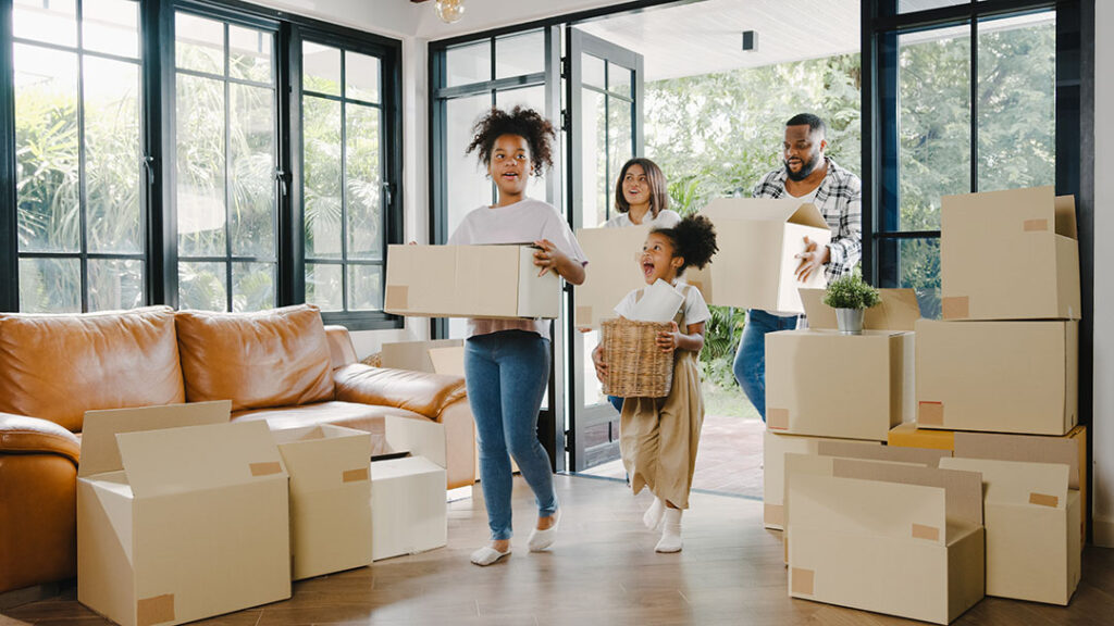 Family moving into a new home each carrying boxes into a large room with floor-to-ceiling windows.